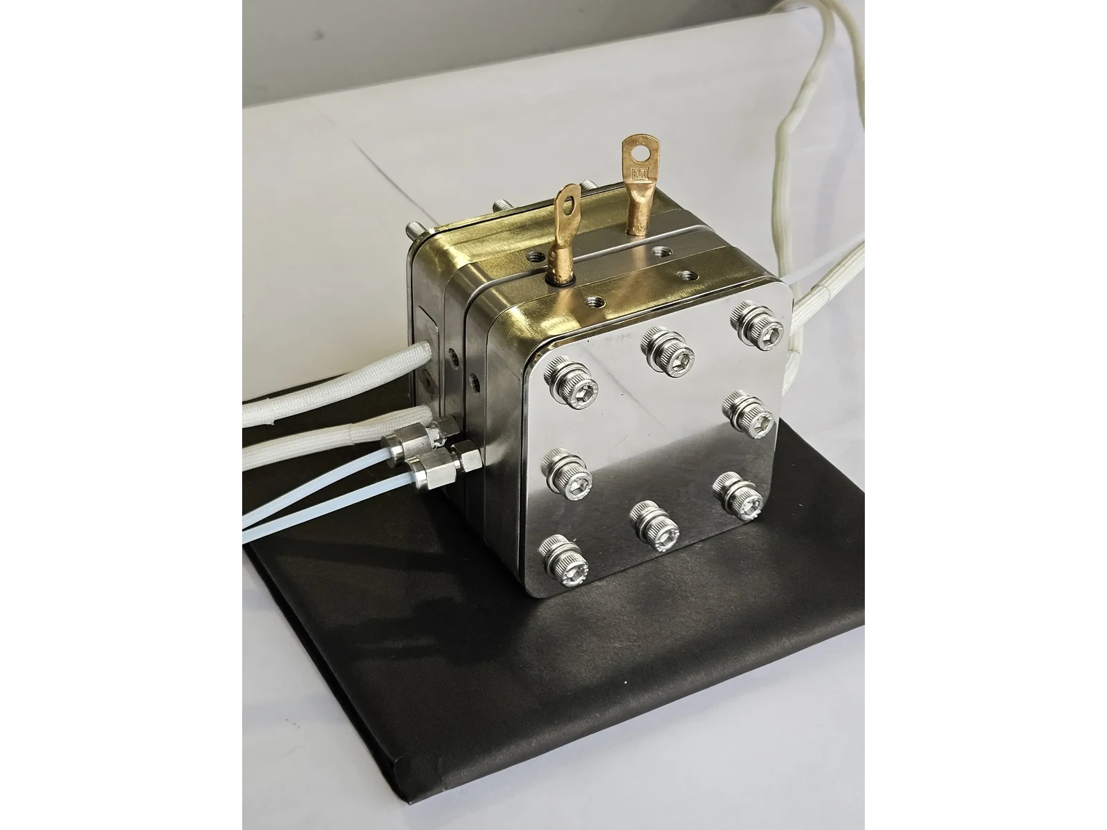High Temperature PEM/AEM Electrolyzer Hardware - three-quarter view of the stainless steel cell with its brass current tabs and compression bolts, standing on a black mat