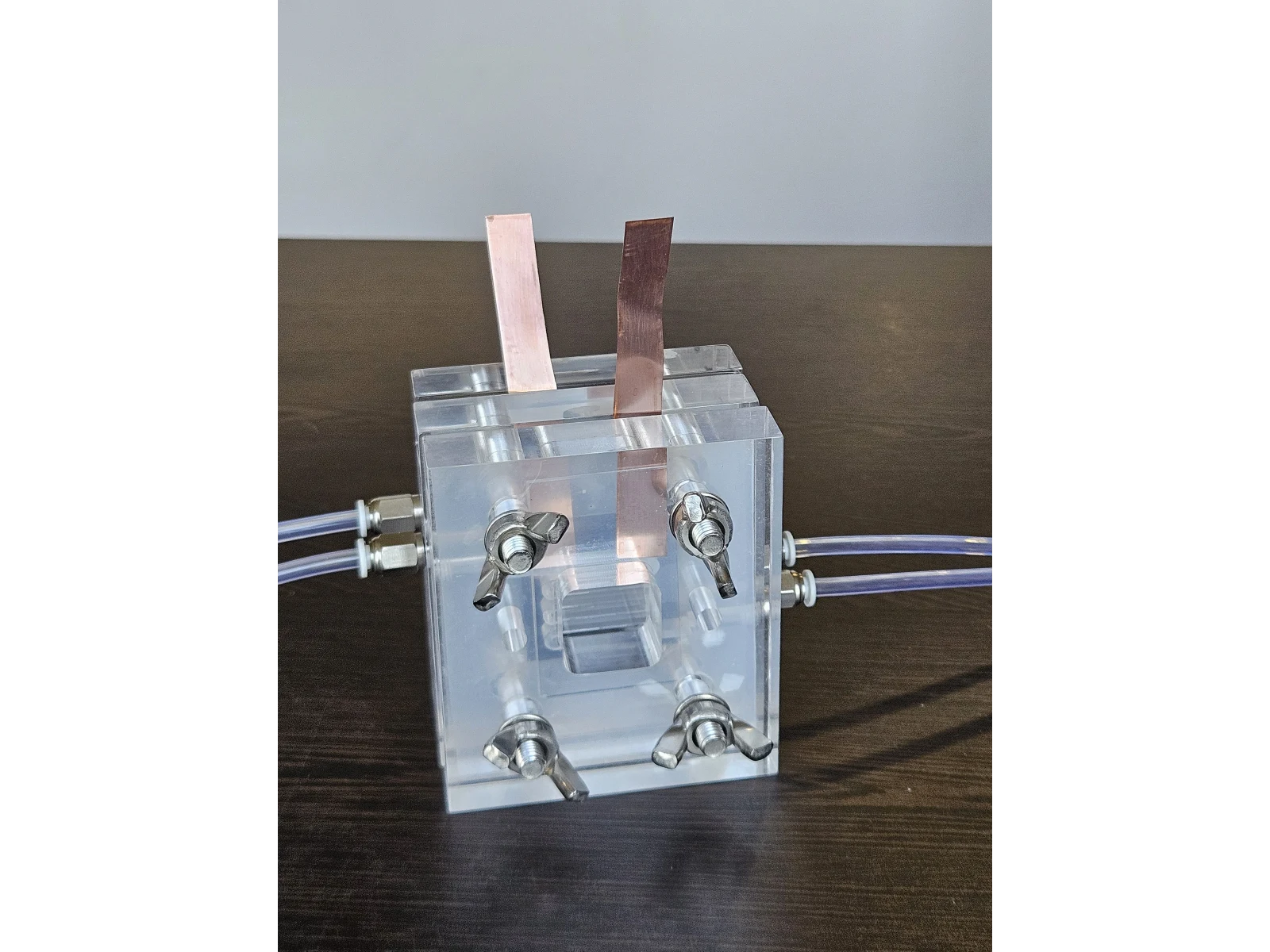 Vanadium Flow Cell Reactor - three-quarter view of the assembled transparent PMMA cell, its two copper current tabs rising from the top, wing screws at the corners and electrolyte tubing entering each side